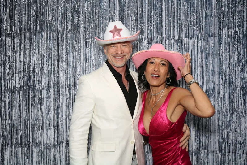 Two guests wearing hats and posing together in front of a silver sequin backdrop