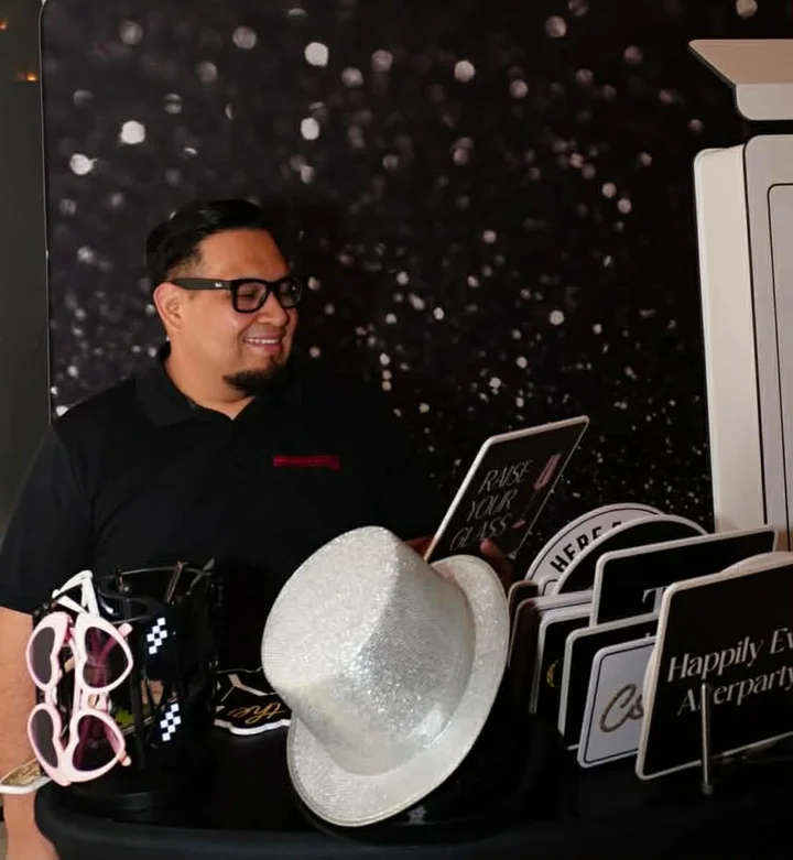 Juan sonríe junto a una cabina de fotos y una mesa con accesorios para bodas