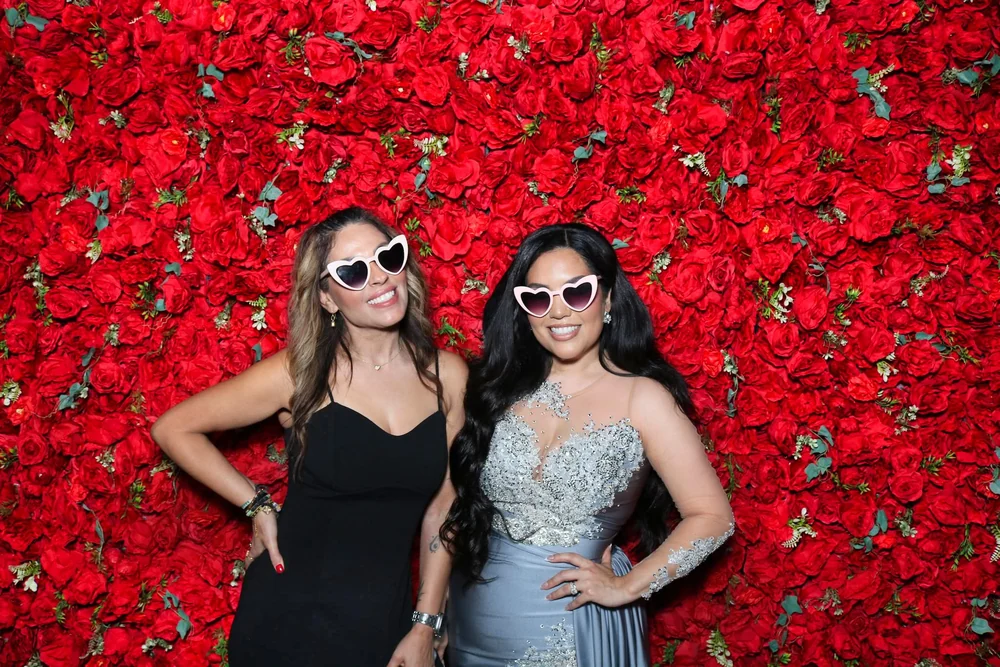 Invitados con lentes de corazón frente a la pared de rosas rojas