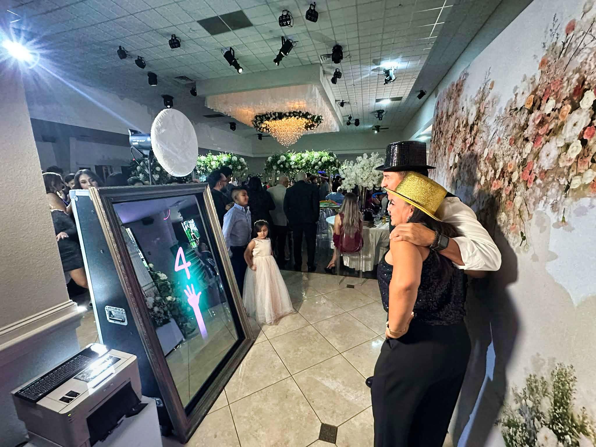 Invitado con sombrero dorado junto a una cabina de espejo enmarcada y un muro de flores